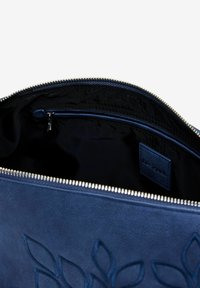 Leather bag in navy blue with embossed leaf design. Interior features a black lining and a zippered pocket with a logo tag. Silver zipper closure.