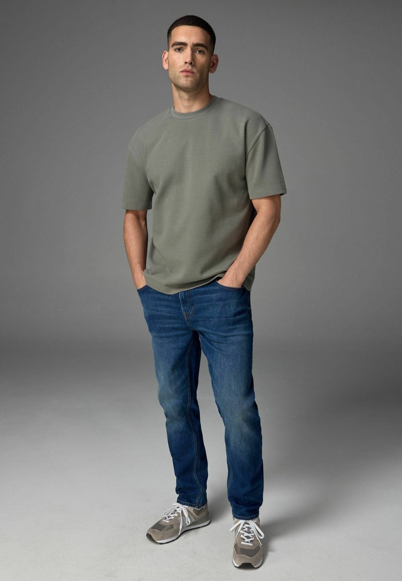 Olijfgroene korte mouwen t-shirt, blauwe spijkerbroek en grijze sneakers met witte veters. De achtergrond is simpel grijs, waardoor de focus op de outfit ligt.