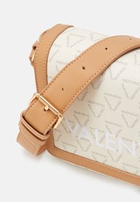 Cinturino in pelle beige con accessori dorati attaccato a una borsa color crema con un delicato motivo logo ripetuto. Texture liscia e forma strutturata.