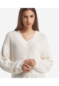 Hvid oversized striktrøje med V-hals, ribbede manchetter og tekstureret mønster. Stoffet ser blødt og afslappet ud.
