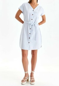 Abito camicia a righe blu chiaro con maniche corte, frontale con bottoni, vita con cintura e aperture laterali; abbinato a sandali bianchi con tacco.