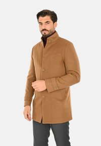 Cappotto in misto lana marrone con colletto alla mandarino, taschino sul petto, chiusure a bottoni e vestibilità su misura. Indossato sopra un dolcevita bordeaux.