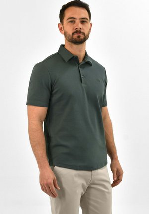 Mann trägt dunkelgrünes kurzärmeliges Polohemd und hellbeige Hose, steht vor einfarbig weißem Hintergrund.