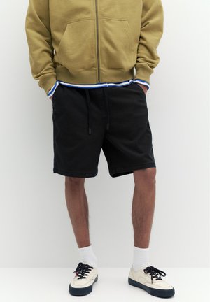 Person iført en beige hættetrøje med lynlås, sorte shorts med snøre, hvide ankelsokker og cremefarvede sneakers med sorte såler og snørebånd.