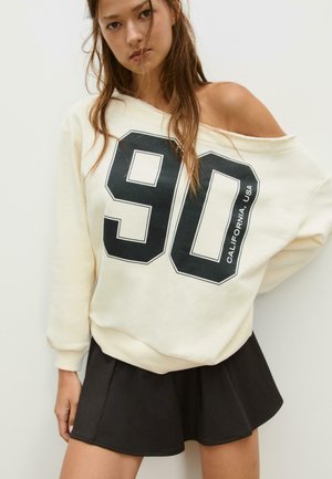 Giovane donna con felpa color crema scollata sulle spalle, con grande stampa "90 California, USA" e gonna mini plissettata nera.