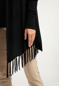 Poncho noir en tricot avec des points texturés, doté d'une longue frange noire. La main du mannequin repose délicatement sur le bord, associé à un pantalon beige.