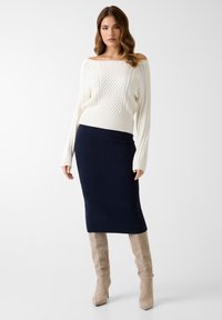 Pull blanc à col bateau en maille torsadée, jupe midi côtelée bleu marine, et bottes ajustées beige jusqu'aux genoux. Textures lisses et couleurs contrastées.