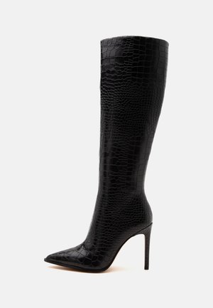 Botte noire haute jusqu'au genou à talon aiguille, avec un bout pointu et une tige en cuir texturé à motif croco sur un fond clair uni.