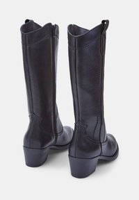 Botas de cuero negro hasta la rodilla con punta redonda, tacón cónico y costuras visibles. Cremalleras laterales que mejoran la accesibilidad.