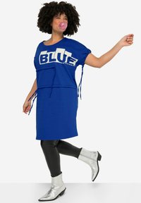 Blauwe oversized jurk met witte grafische tekst, aangesnoerde zijkanten, gedragen over zwarte leggings en zilveren enkelboots met puntige neuzen.