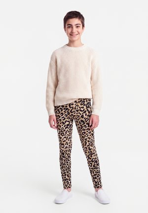 Jeune personne souriante, portant un pull en maille crème, des leggings à motif léopard et des chaussures blanches sans lacets, debout sur un fond blanc.