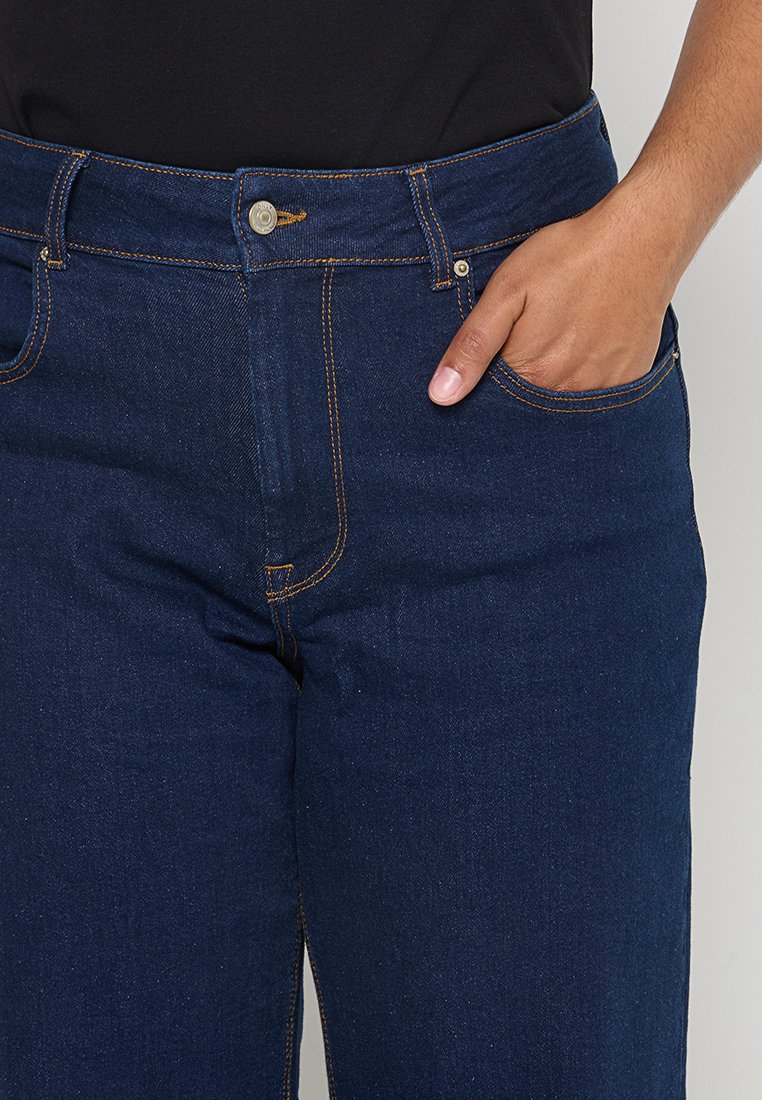 Jean en denim bleu foncé avec une taille ajustée, des détails de couture subtils et des poches latérales. Le tissu a une texture lisse.