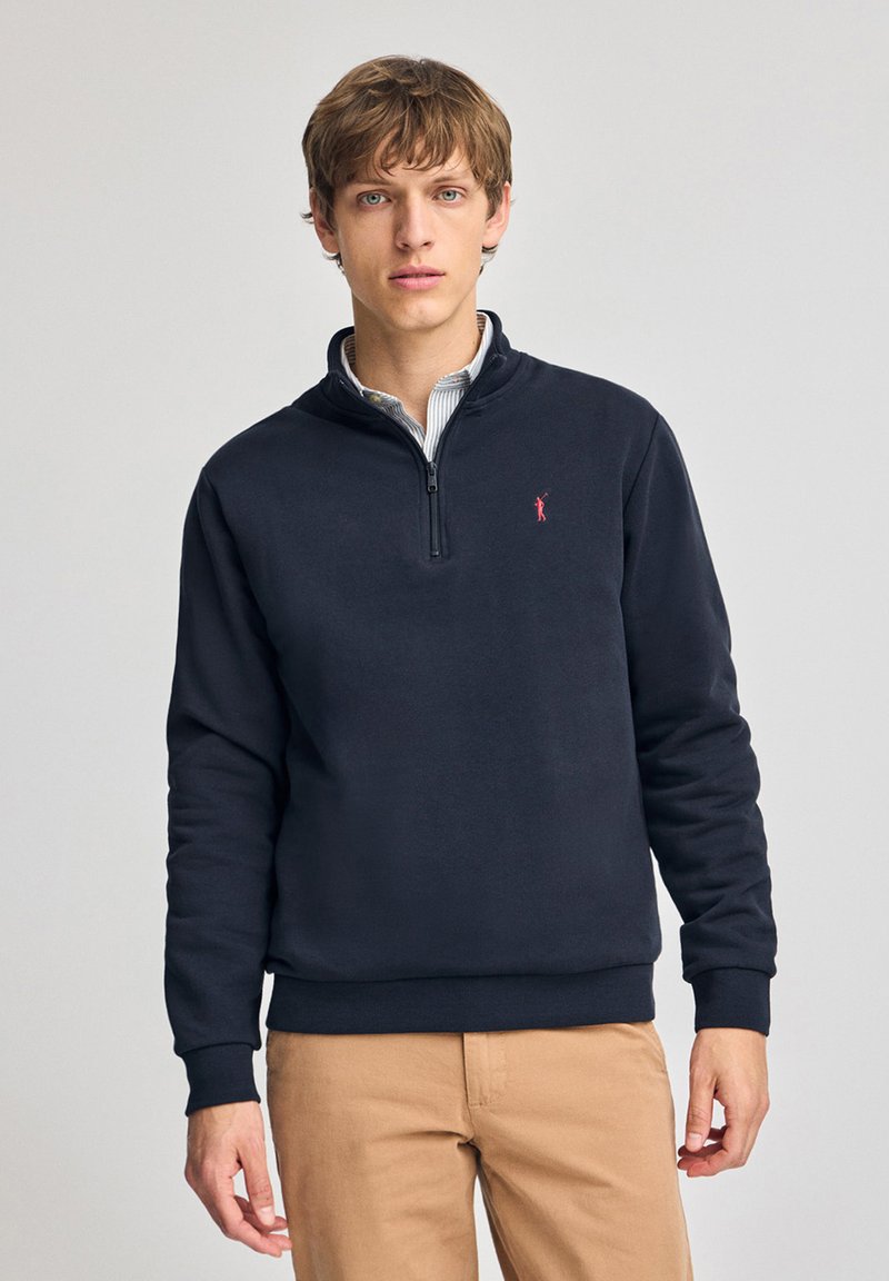 Marine blauwe kwart-rits sweatshirt van zachte stof, voorzien van een rood logo op de borst, geribbelde manchetten en onderrand. Draagt over een geblokte blouse.