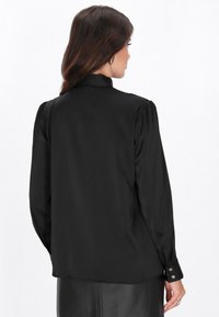 Hosszú ujjú fekete blúz sima anyagból, magas gallérral, pliszírozott ujjakkal és arany gombos díszítéssel a mandzsettákon.