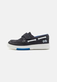 Scarpa in suede blu con cuciture bianche, cinturino in velcro e un accento blu sulla suola; presenta una suola in gomma bianca testurizzata.