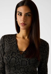 Kobieta o długich ciemnych włosach, ubrana w czarno-szary top z dekoltem w serek, zwrócona twarzą do przodu z neutralnym wyrazem twarzy na jednolitym tle.