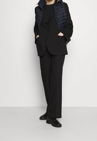 Czarny garnitur na miarę z szerokimi spodniami i oversize'ową marynarką, zestawiony z granatową, pikowaną kamizelką. Gładka tkanina z subtelnymi fakturalnymi detalami.