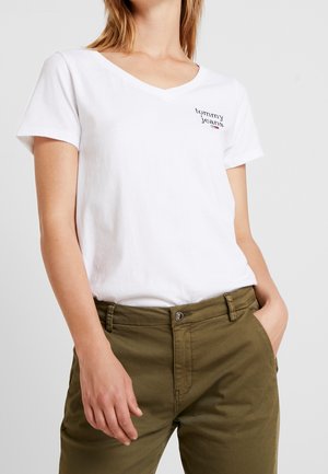 Kobieta w białej koszulce typu V-neck od Tommy Jeans i oliwkowych spodniach, z jedną ręką w kieszeni, a drugą swobodnie opartą przy ciele.