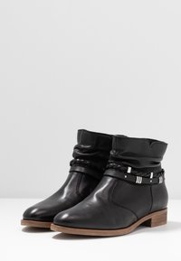 Botas de tobillo de cuero negro con parte superior arrugada, correa trenzada, detalles plateados y tacón bajo de madera. Textura suave y diseño de punta afilada.