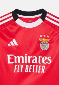 Crvena nogometna majica s crnim i bijelim detaljima, koja sadrži Adidas logo, tekst "Emirates Fly Better" i grb kluba s orlom.