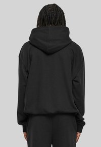 Persona con cabello trenzado oscuro, vistiendo una sudadera negra de gran tamaño y pantalones negros a juego, de espaldas frente a un fondo gris liso.