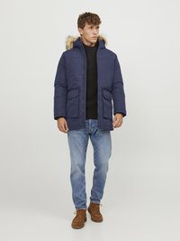 Giacca puffer blu navy con cappuccio bordato di pelliccia, due tasche frontali, abbinata a un maglione nero, jeans blu e scarpe in stile mocassino marroni.