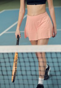 Roze tennisrok met een uitlopende stijl en vochtregulerende stof, gecombineerd met een zwarte crop top en zwarte sportschoenen. Tennisracket wordt vastgehouden.