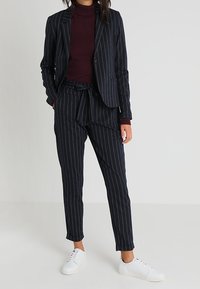 Costume bleu marine à rayures avec une veste ajustée et des pantalons fuselés ; associé à un col roulé bordeaux et des baskets blanches.