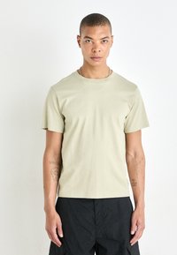 Svetlo olivnozeleni bombažni t-shirt s krojem, kratkimi rokavi in prilegajočo se silhueto, prikazan na osebi proti nevtralni ozadju.