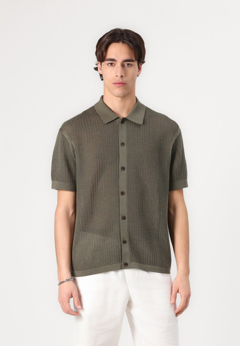 Polo shirt tricotat verde cu mâneci scurte, cu nasturi pe față și o textură moale. Are un guler clasic și nasturi maro.