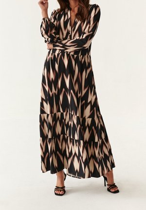 Sukienka maxi z czarno-beżowym wzorem w zygzaki, długimi rękawami, głębokim dekoltem oraz warstwowymi falbanami przy brzegu. Noszona z czarnymi szpilkami.