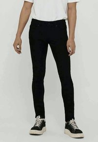 Melnas šauras džinsu bikses, kas izgatavotas no denima, ar klasisko piecu kabatu dizainu, smalku šuvi un gludu apdari. Kombinētas ar melniem sporta apaviem.