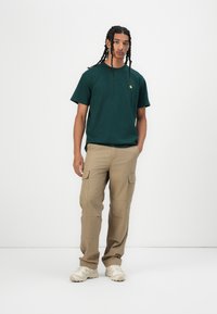 Green cotton t-shirt with a small yellow logo, paired with beige cargo pants featuring side pockets and a relaxed fit. White sneakers.