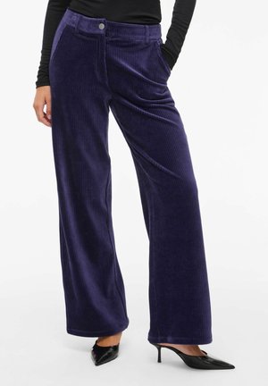 Vrouw draagt donkerpaarse wijde corduroy broek met steekzakken aan de voorkant en zwarte pumps met spits toelopende neus en kleine hak, hand in zak.