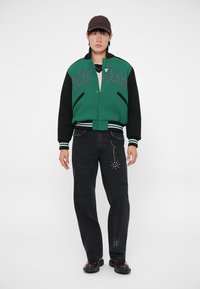 Giacca varsity verde e nera con design a contrasto, bottoni a pressione, orlo a righe, abbinata a jeans neri decorati con dettagli in borchie.