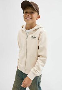 Beżowa bluza z kapturem na zamek z przednią kieszenią i logo JJ Studios. Noszona na białej koszulce, zestawiona z zielonymi dżinsami i brązową czapką.