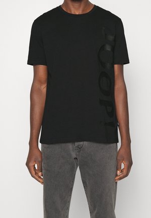 T-shirt en coton noir avec un col rond, des manches courtes et un design graphique textuel tonal sur le côté gauche. Texture douce, coupe décontractée.