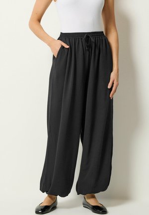 Frau trägt schwarze, weit geschnittene Hosen mit elastischem Bund und Kordelzug, kombiniert mit schwarzen Ballerinas und weißem ärmellosem Oberteil.