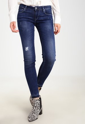 Kobieta ubrana w ciemnoniebieskie obcisłe jeansy z delikatnym przecieraniem i botki na koturnie w panterkę, stojąca na tle jednolitego tła.