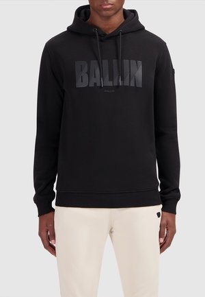 Homme portant un sweat à capuche noir avec le texte « BALLIN » et un pantalon beige, sur un fond clair uni, faisant face à l'avant.