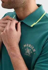 Tyrkysová polo košeľa so štruktúrovaným povrchom, s golierom s žltými akcentmi a logom na hrudi s nápisom "MUNICH 1939."