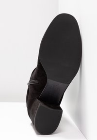 Bottine en daim noir avec fermeture éclair, montrant une semelle lisse et un talon texturé, placée contre un mur d'angle blanc.