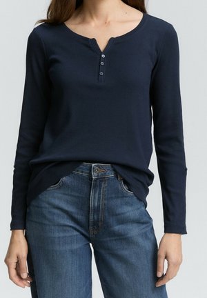 Woman wearing a navy blue long-sleeve henley shirt with three buttons and blue denim jeans, standing with hands relaxed by sides.