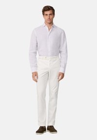 Mann in einem helllila, knöpfbaren Hemd, weißen Hosen und braunen Slippern, der mit dem Gesicht nach vorne vor einem einfarbigen Hintergrund steht.