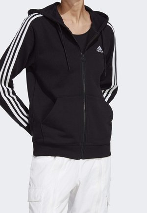 Persona con sudadera con capucha negra Adidas con cremallera, con rayas blancas en las mangas y logo, combinada con pantalones cargo blancos.