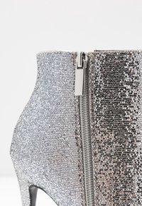 Bota de tacón alto plateada con lentejuelas y detalle de cremallera. La superficie presenta un patrón texturizado y brillante, y la forma es elegante y afilada.