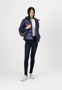 Marineblå pufferjakke med hette, glidelås og quiltet tekstur. Kombinert med en hvit topp, mørke jeans og en grå skulderveske.
