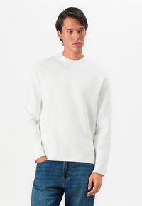 Hombre vestido con un suéter blanco de manga larga y jeans azules, de pie con una mano en el bolsillo, mirando ligeramente hacia un lado contra un fondo blanco.