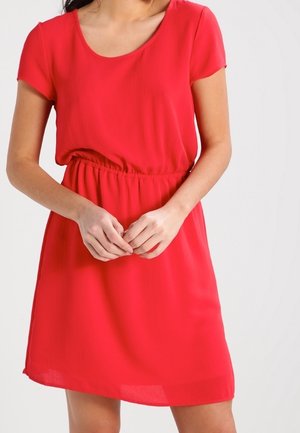 Femme portant une robe rouge à manches courtes avec un col rond et une taille élastique, les mains légèrement jointes devant elle.