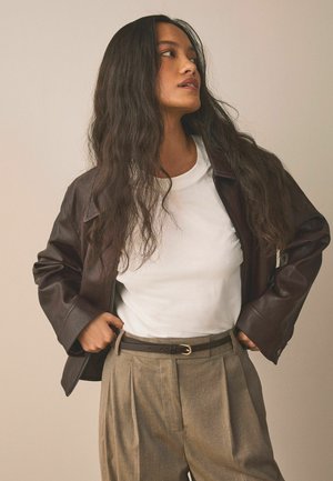 Braune Lederjacke, weißes T-Shirt und beige plissierte Hose mit braunem Gürtel. Glatte Textur, lässiges Design und neutrale Farbpalette.
